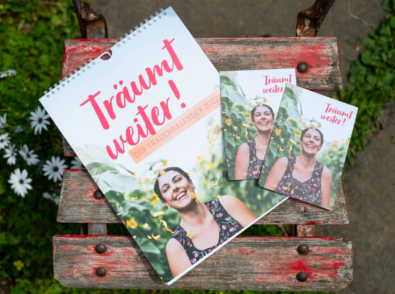 Titelbild des Frauenkalender mit dem Titel "Träum weiter" in rosa Schreibschrift und einer lachenden Frau im Sonnenblumenfeld.